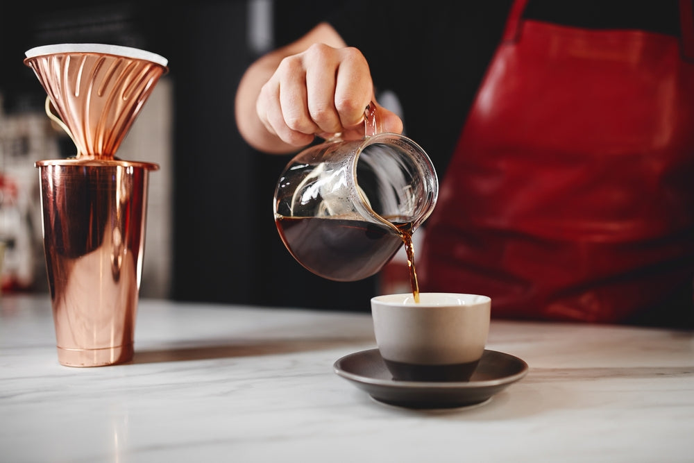 Barista brewed specialty coffee in a v60 brewer and then pours coffee from glass kettle in cup.
