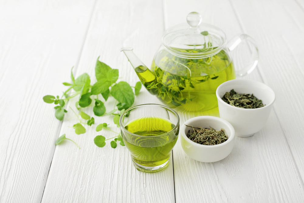 Green tea brewed in a glass teapot and tea cup with green tea leaves in white bowls.